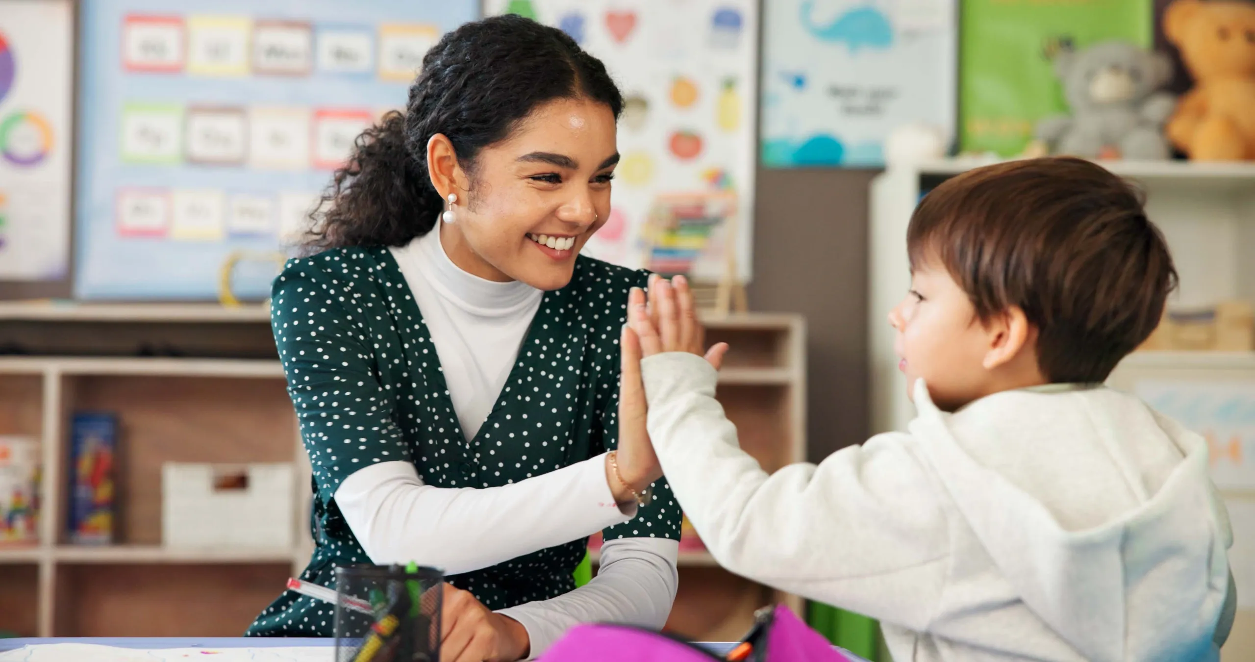 A teacher giving a young boy a high-five at his desk in a colorful classroom